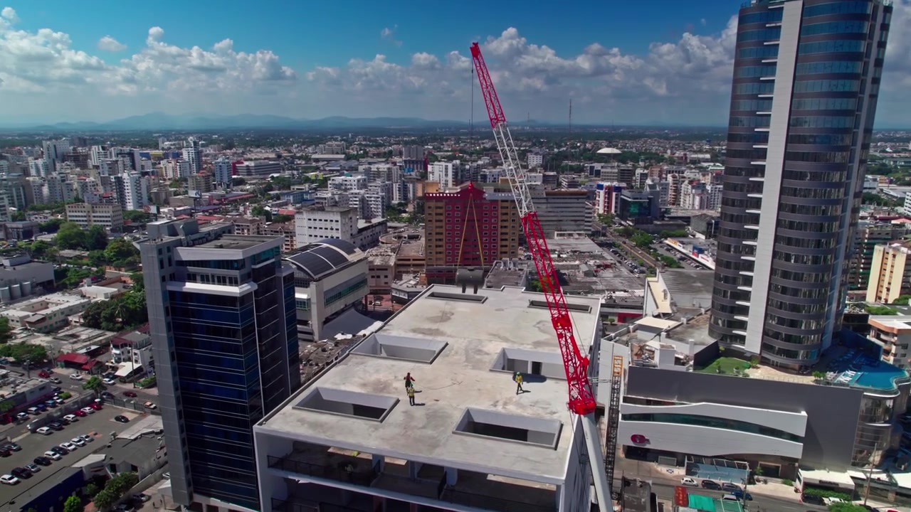 Toma aérea de dron en picado orbitando a la izquierda alrededor de un edificio en construcción con una grúa móvil roja, revelando modernas torres comerciales y el horizonte de la ciudad en Santo Domingo.