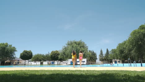 two women in swimwear walking towards a swimming pool on a sunny day