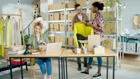 equipo de diseñadores multiétnicos escribiendo en tabletas y casillas de verificación, empacando ropa y escaneando paquetes en una tienda de ropa de moda