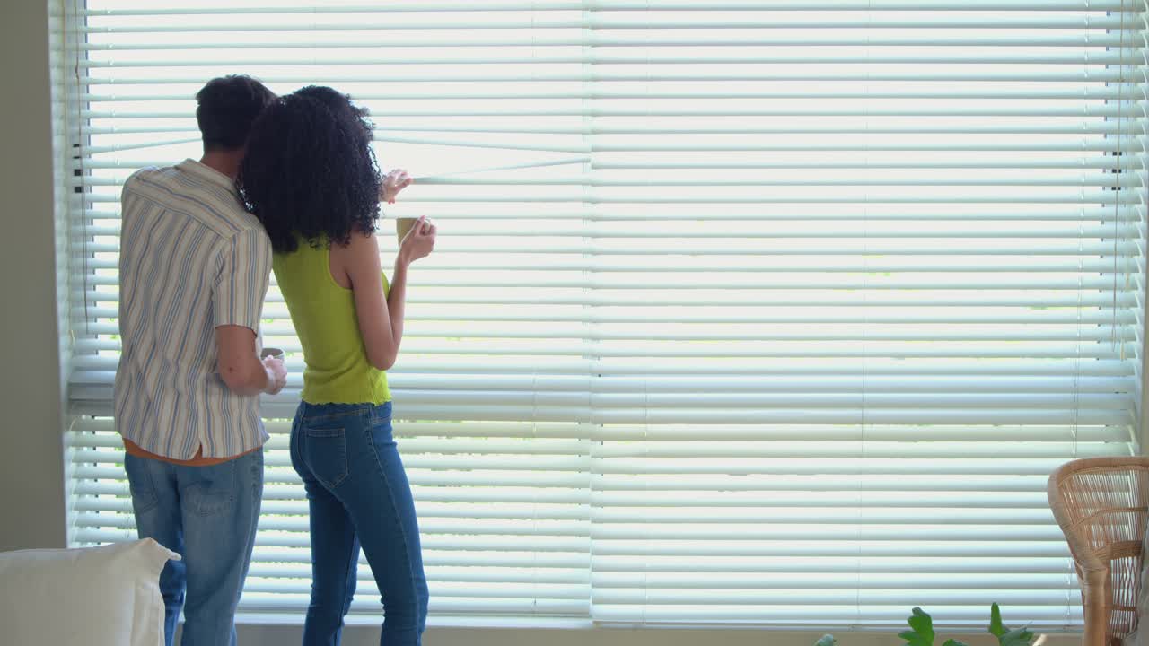 Diverse couple in living room, man approaching, adjusting blinds via cord while embracing