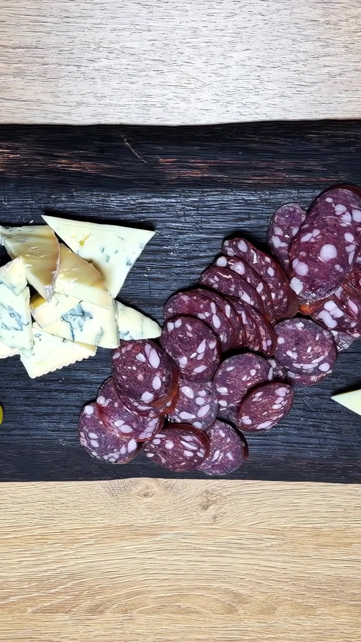 Salsiccia stagionata affettata e spicchi di formaggio erborinato disposti su un tagliere di legno scuro, su una superficie di tavolo in legno chiaro. Video verticale, ideale per i social media.