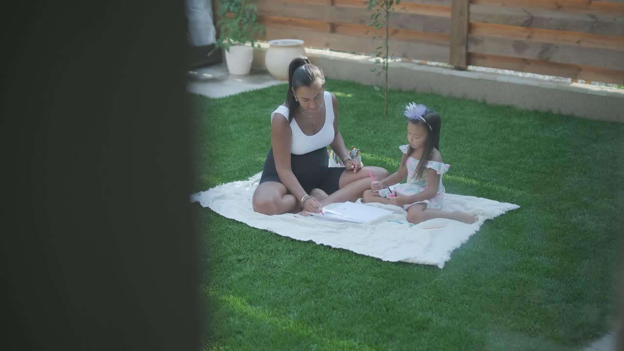 A mother and her young daughter sit on a blanket in the backyard, engaging in a drawing activity together. The outdoor scene captures a peaceful family moment, fostering creativity and bonding.
