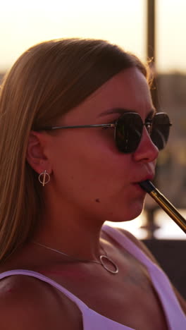 blonde girl in sunglasses takes a pipe of a hookah to her lips. woman makes smoke into air in sun rays. blurred backdrop. vertical video