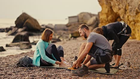 A-blonde-girl-in-a-black-sports-summer-uniform,-a-blonde-girl-with-a-bob-hairstyle-and-a-brunette-guy-in-a-gray-T-shirt-put-on-special-climbing-shoes-and-prepare-to-climb-yellow-rocks-on-a-stony-and-rocky-seashore-in-the-summer