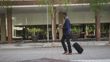 African-american-businessman-with-face-mask-walking-with-suitcase