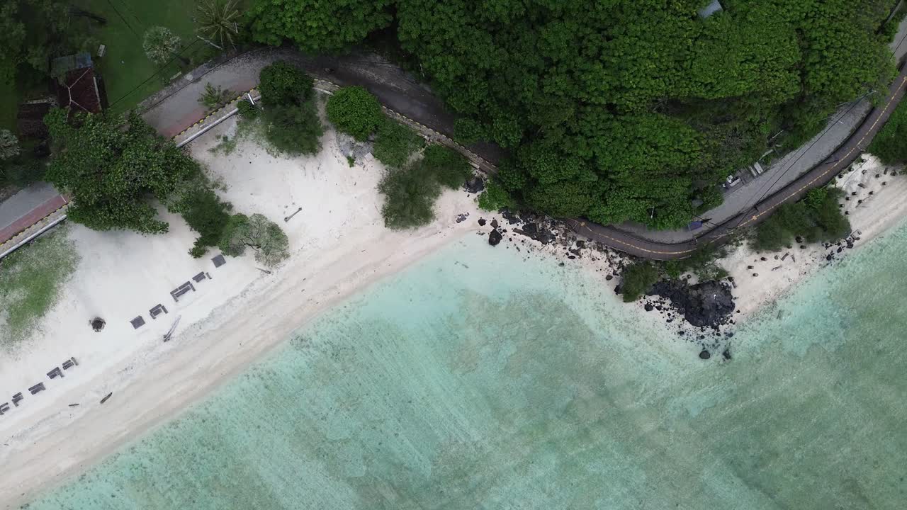 Static top-down aerial image showing white sand, sunbeds and lush trees. Clear water outlines the shoreline under soft light