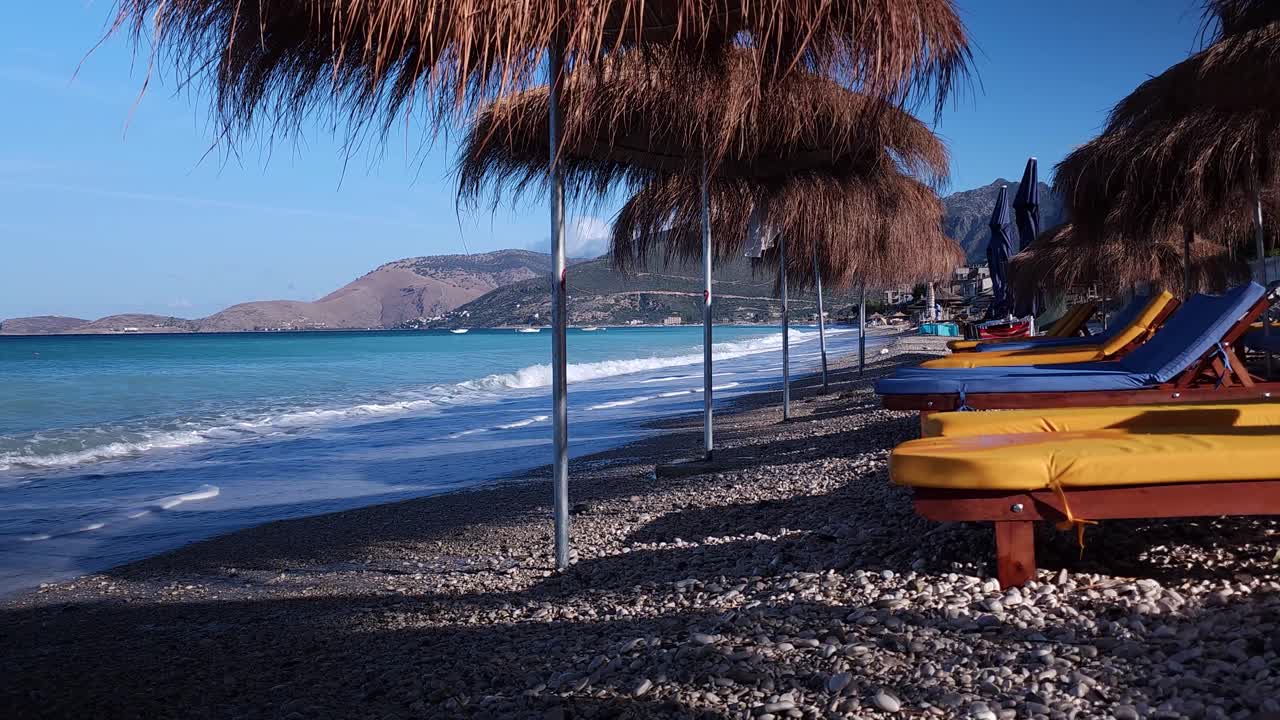 Peaceful beach with straw umbrellas and sunchairs in Albania, turquoise sea water and white waves washing pebbles, summer vacation background