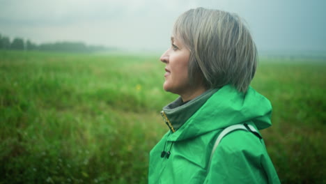 vista lateral de una mujer mayor con un impermeable verde, caminando por un camino rodeado de vegetación, con una expresión enfocada en su rostro bajo un cielo suave y nublado, se mueve con calma