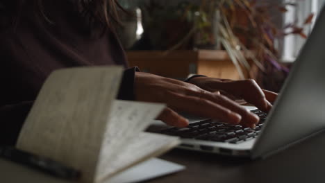 an asian lady types on her laptop keyboard with a notebook containing notes is on the desk next to her