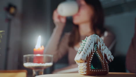 close-up of red candle burning beside detailed gingerbread house with white icing, woman in background drinks tea, slightly blurred, with soft warm lighting