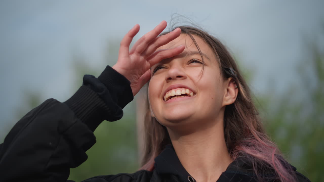 Niña sonriendo bajo suaves gotas de lluvia cerca de un charco, adolescente alegre disfrutando de una ducha de verano, cabello mojado con reflejos rosados, chaqueta negra, fondo de árboles verdes, primer plano espontáneo, expresión esperanzadora