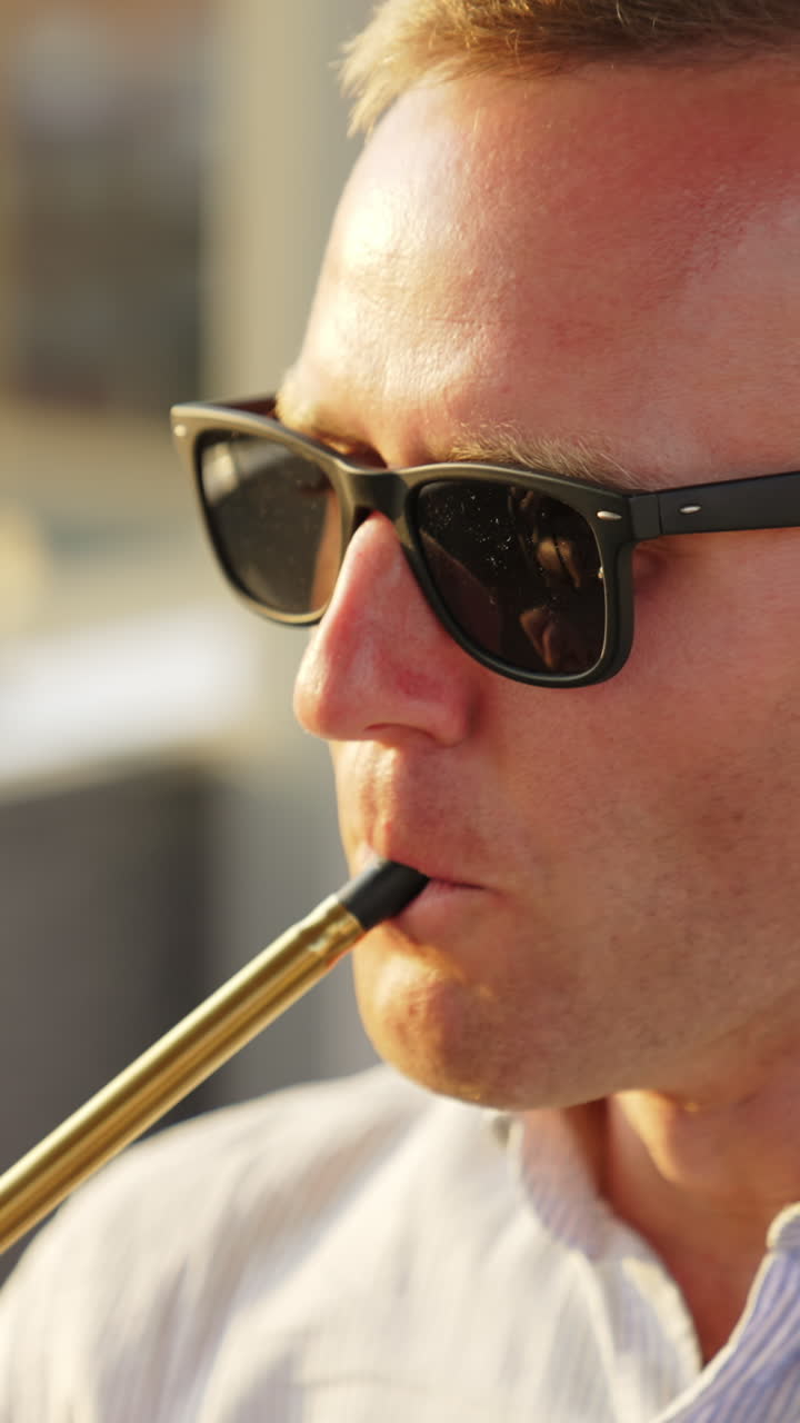 Mid-aged man in sunglasses smoking chillum sitting outdoors. Chilling male rises head and breathes out white smoke. Close up. Vertical video