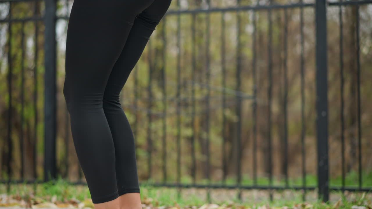 Close-up view of woman stretching her legs outdoors in black leggings and athletic wear, focus on leg movements during stretching in park, highlighting fitness routine and healthy lifestyle