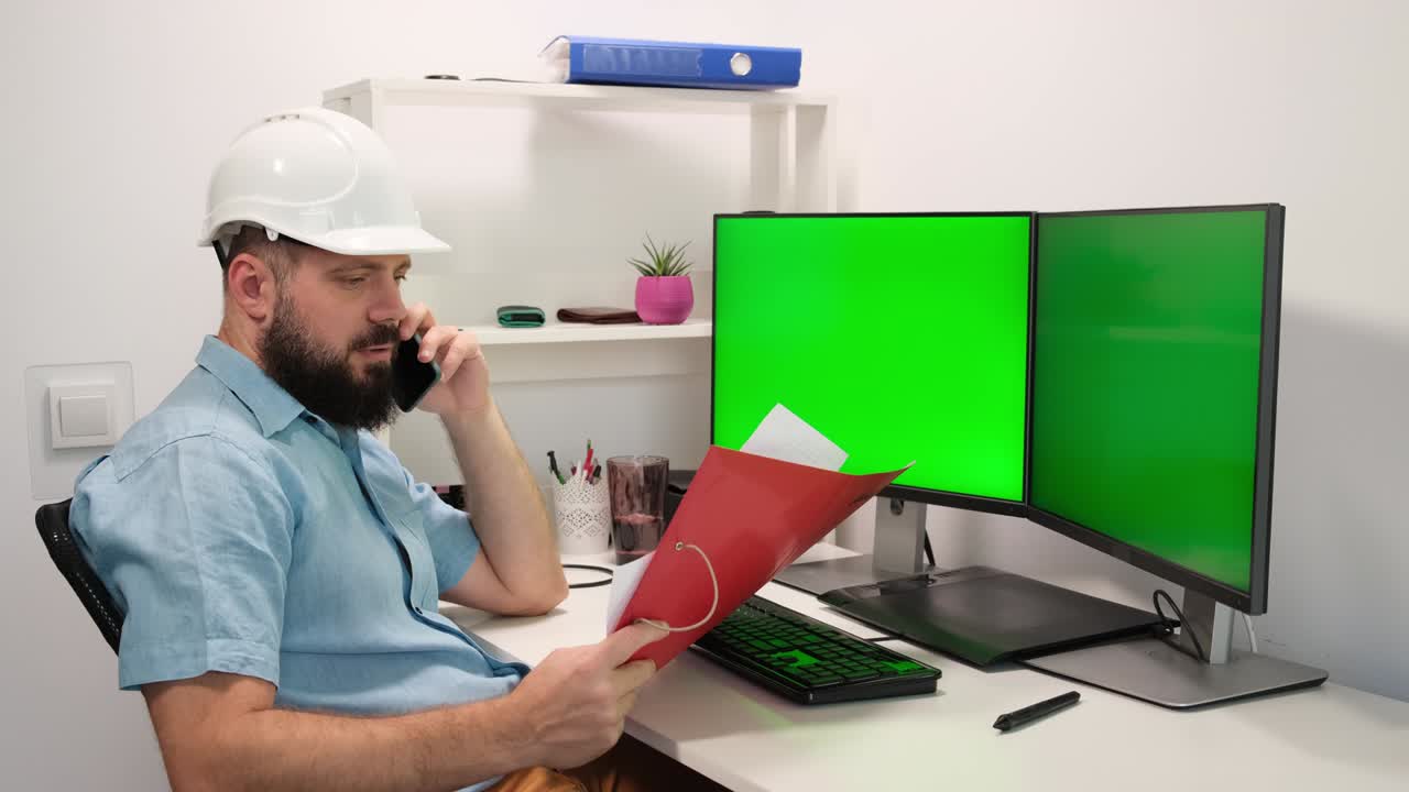 ingeniero senior en casco está trabajando en una computadora de escritorio con dos pantallas verdes en los monitores y hablando por teléfono en un gabinete.
