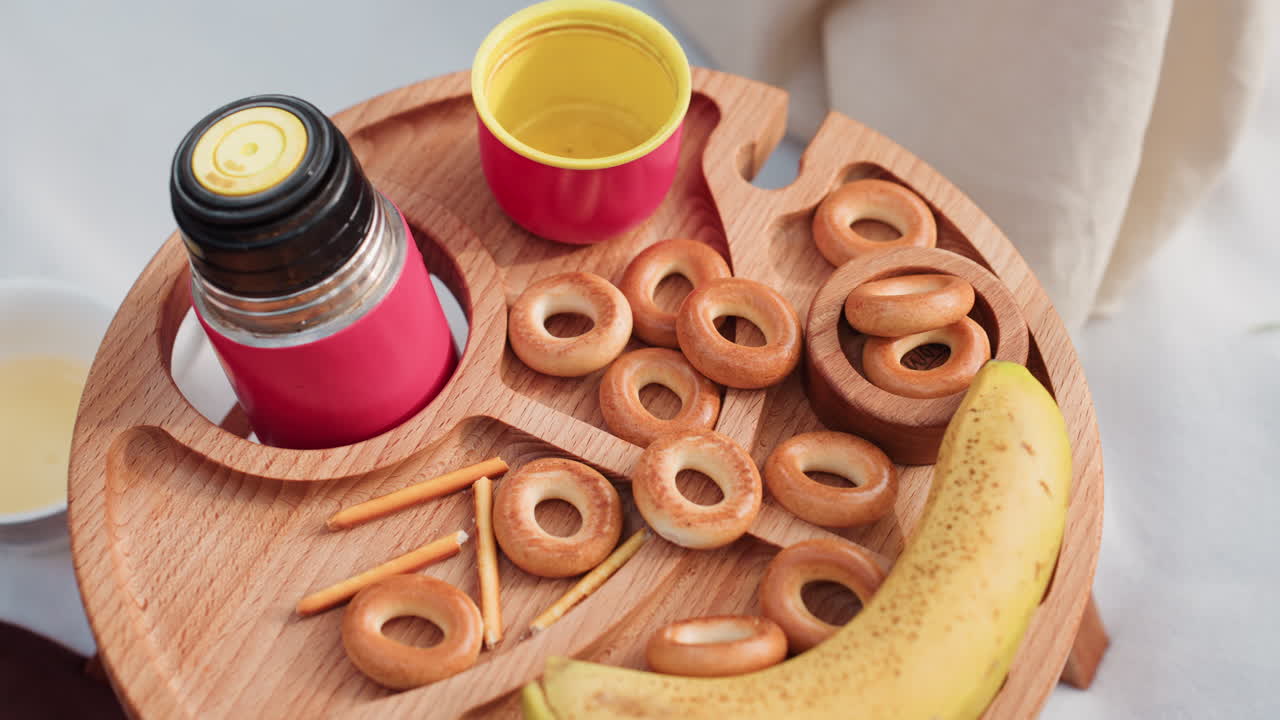 따뜻한 햇살 아래 야외 피크닉을 준비하는 동안 바나나, 베이글, 보온병, 브레드스틱이 담긴 나무 쟁반이 놓인 작은 피크닉 테이블의 항공 뷰에서 손이 뻗어 베이글 하나를 집으려고 하고 있어요.