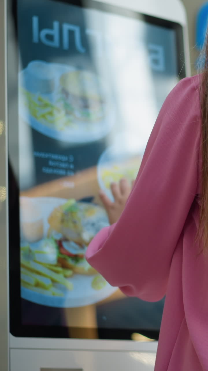 vista trasera de una dama en vestido rosa con un bolso negro interactuando con una máquina de pedidos de alimentos de pantalla táctil en un centro comercial bien iluminado, se puede ver un ligero borrón de personas y elementos de fondo