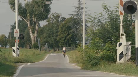 A-bicyclist-is-pedaling-down-a-small-country-road-and-away-from-the-room,-passing-through-a-railroad-crossing-on-a-summer-day