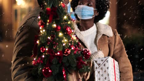 Close-up view of joyful African American couple wearing facial masks talking and holding a Christmas tree while it¬¥s snowing on the street in Christmas