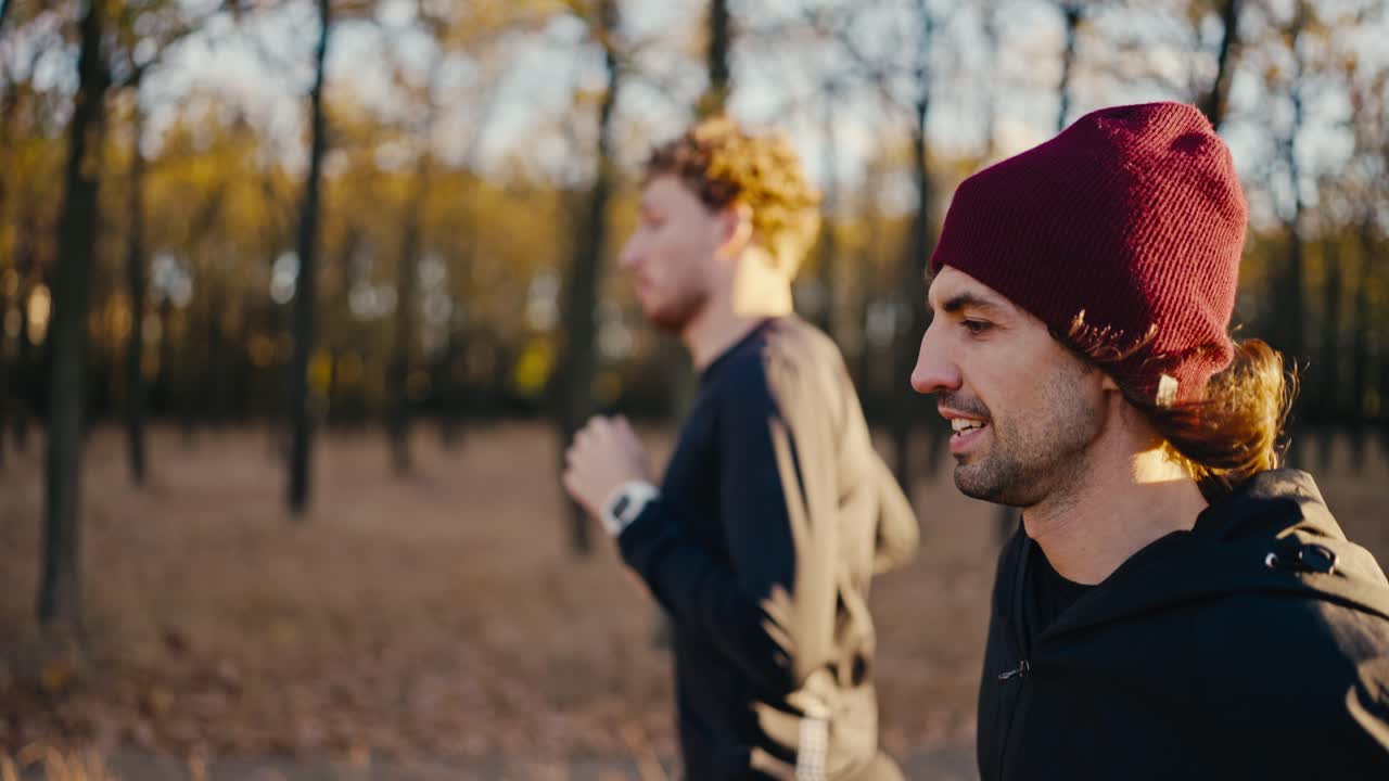 Side view of two brunette guys in black sportswear a guy in a red hat running along an earthen path in the autumn forest in the morning during his jog