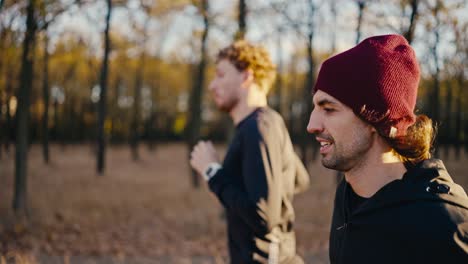 Side-view-of-two-brunette-guys-in-black-sportswear-a-guy-in-a-red-hat-running-along-an-earthen-path-in-the-autumn-forest-in-the-morning-during-his-jog