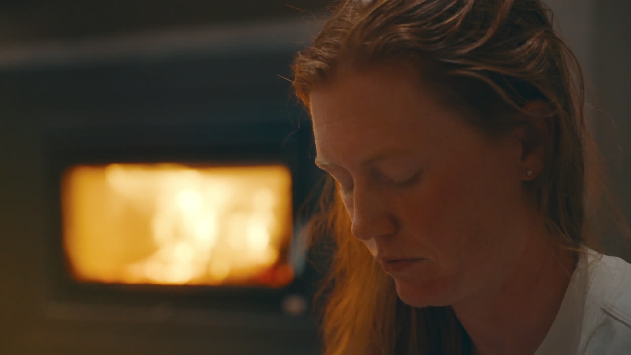 Young redhead woman staring contemplatively with fireplace in background, rack focus to her serious face as she looks up, medium handheld 4k