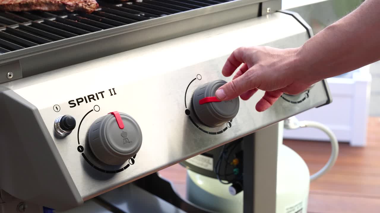 A hand adjusts knobs on a gas grill, controlling heat levels. Bright lighting highlights the stainless steel surface and cooking area