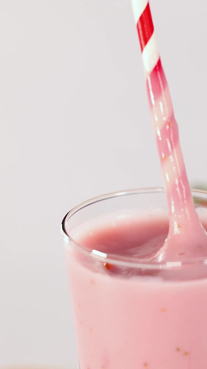 Inquadratura verticale ravvicinata di una mano che mescola un denso frullato rosa con una cannuccia rossa e bianca.
