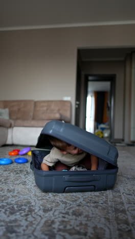lovely baby boy sits in the big grey suitcase. cute kid gets out of suitcase. toys on the floor at backdrop. vertical video.