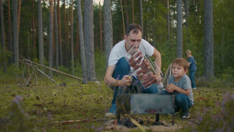 man is grilling sausages for picnic at nature showing to his little son family rest at weekend at summer