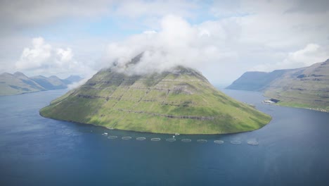 a lush green mountain island surrounded by a serene fjord and fish farming pens in klaksvik, faroe islands. clouds drift along the summit, creating a tranquil and picturesque scene