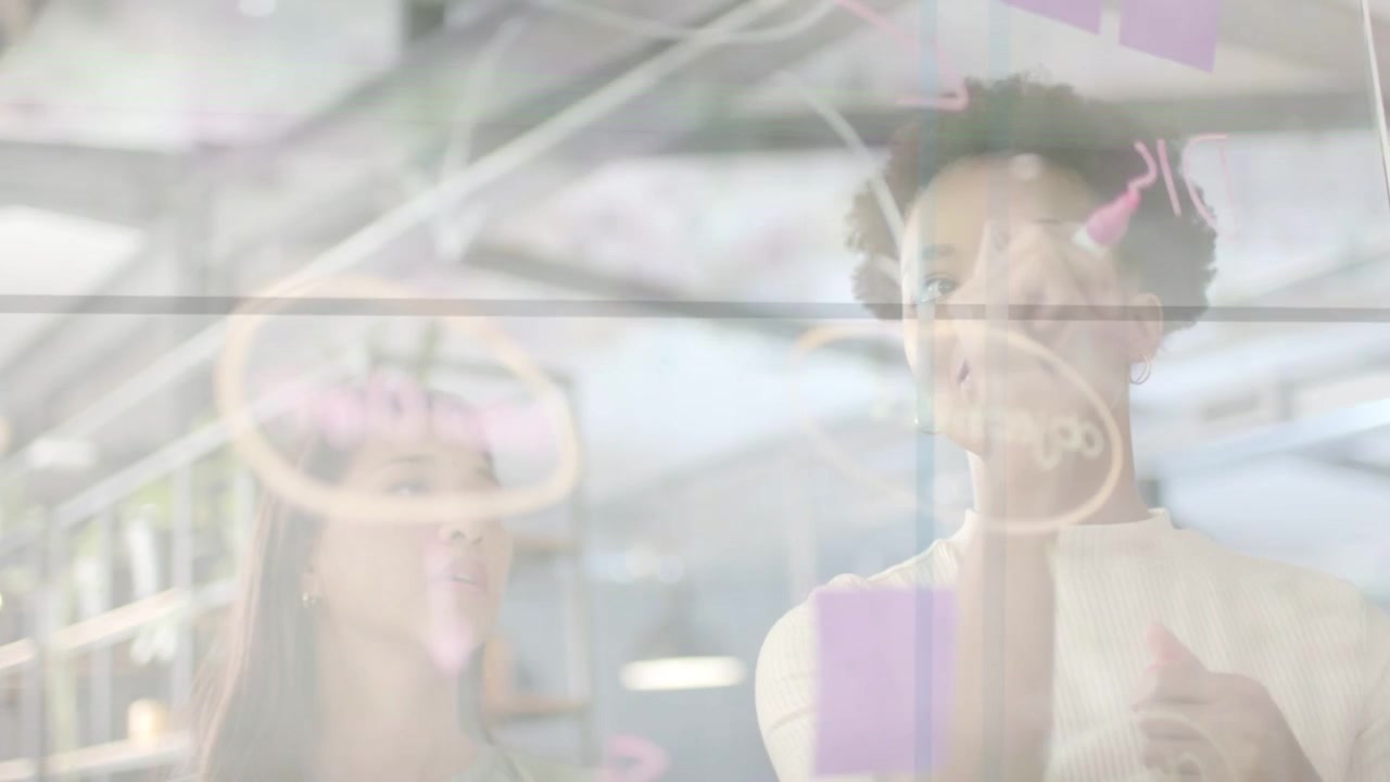 Right woman grabbing pink marker, drawing on glass wall for business planning, colleague observing