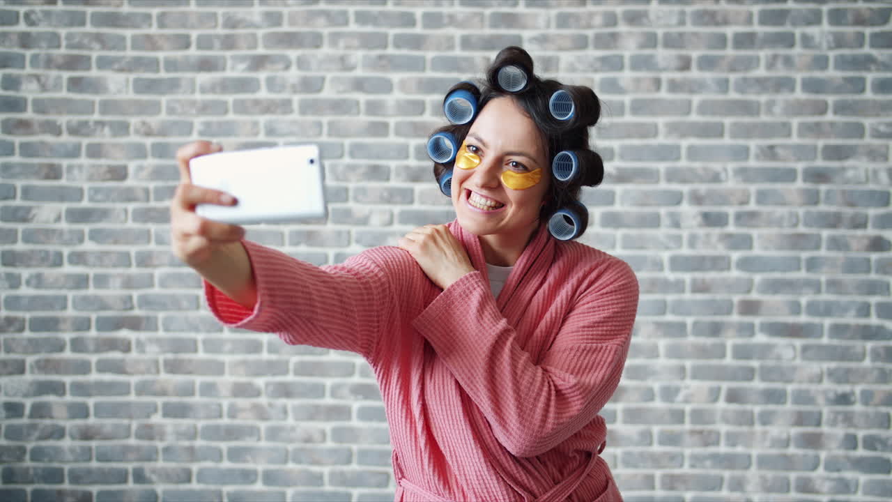 Woman Taking a Funny Selfie in a Bathrobe with Facial Mask and Hair Curlers