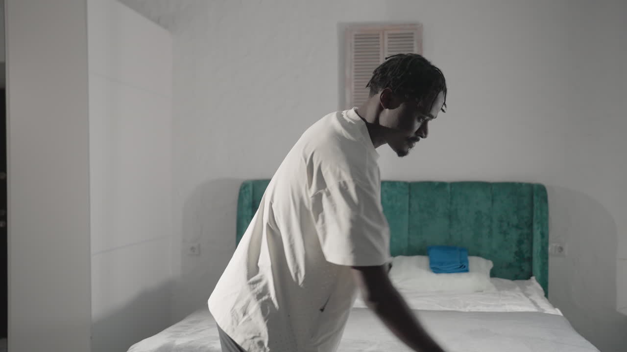 Man in white t-shirt holding backpack, walking through minimalist bedroom with green headboard, sunlight pouring in through the window, preparing to leave home in a clean, simple setting