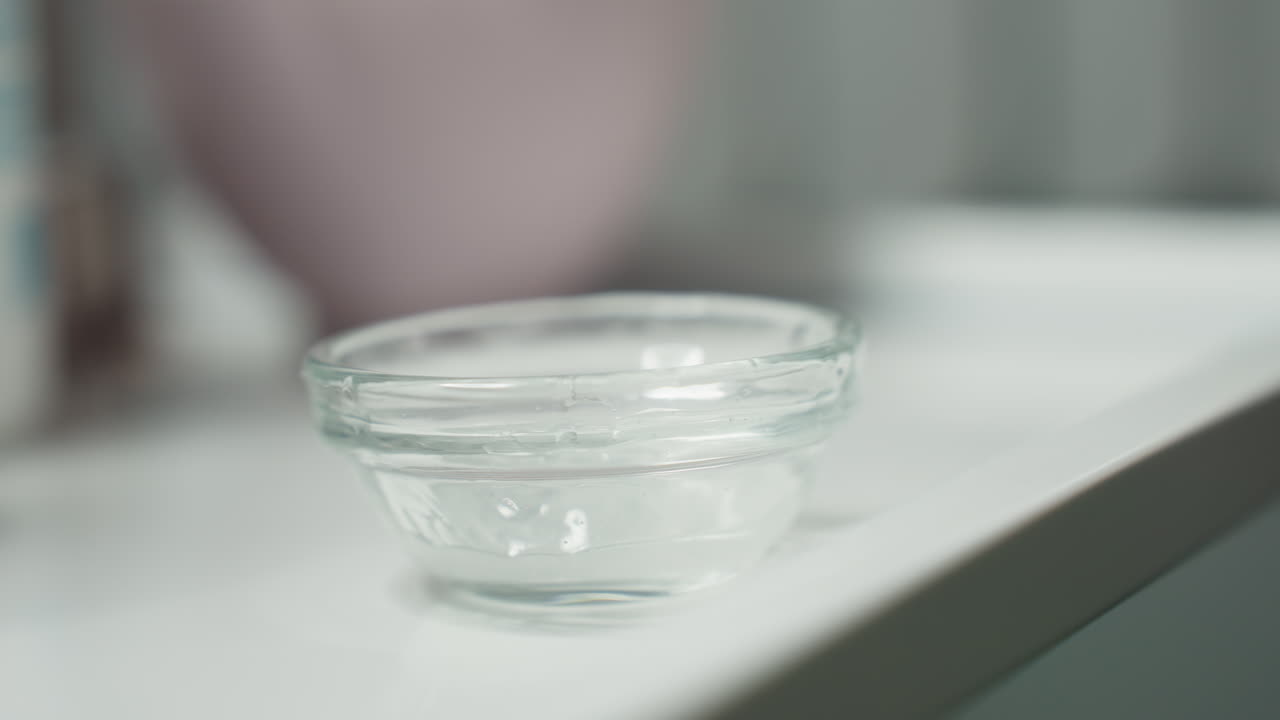 Small glass bowl placed on white tray as gloved hand pours clear viscous gel from bottle tip into bowl under soft spa lighting close up of skincare prep ritual showcasing gel clarity