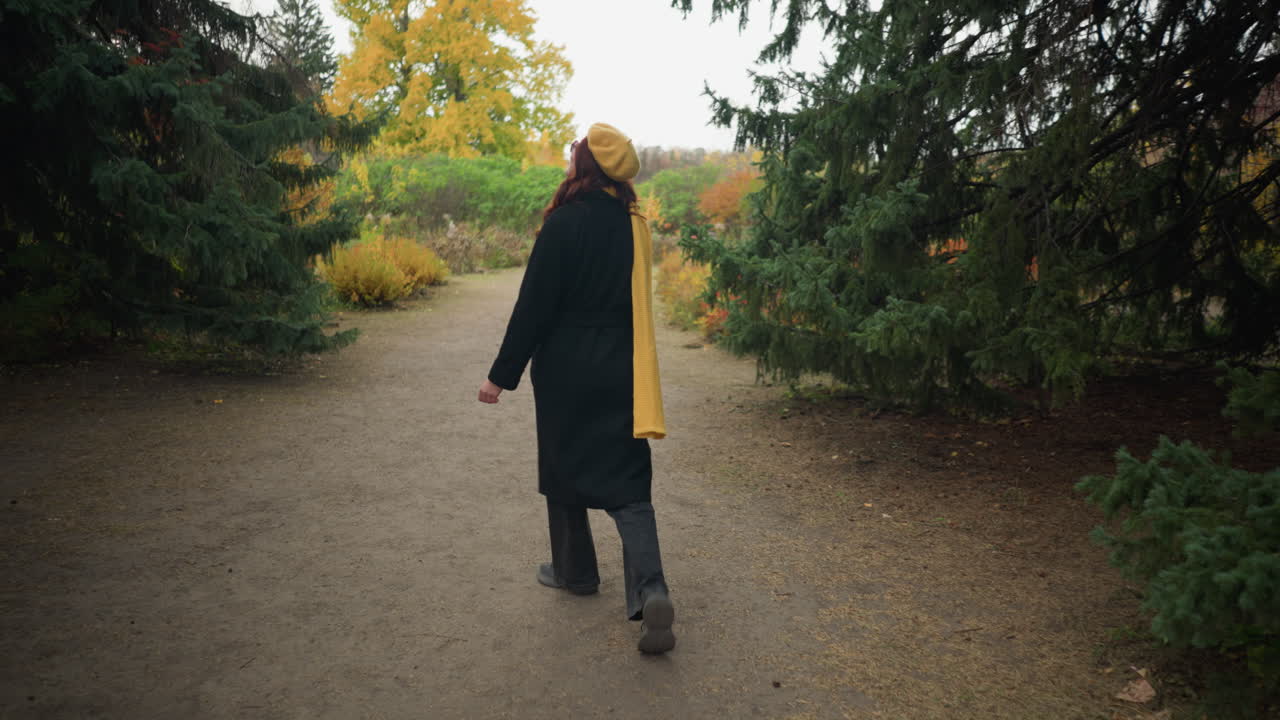 긴 은 머리카락과 노란색 베레트, 스카프, 선글라스를 입은 즐거운 여인이 가을 잎자루로 된 정원에서 걸어가는 동안 행복하게 돌아서 가을의 본질을 구현합니다.