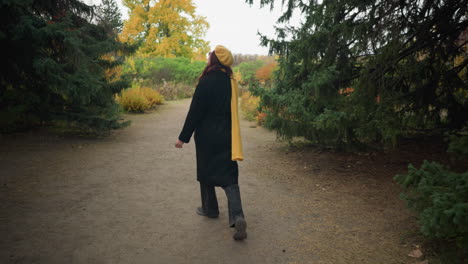 joyful lady with long red hair, wearing a yellow beret, scarf, and sunglasses, happily turning around as she walks in a garden with autumn foliage, embodying the essence of fall