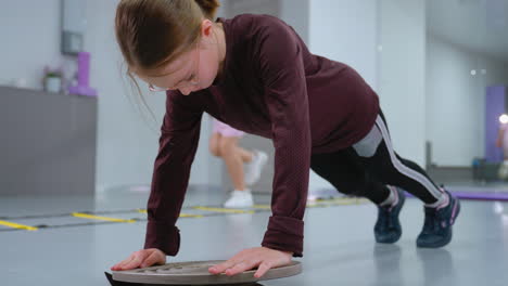 young girl performing stability exercise using balance board to improve strength and coordination while another child jumps in background with mirror reflecting gym surroundings and workout equipment