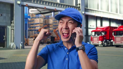 Close Up Of Asian male Courier In Blue Uniform talking On Smartphone And Celebrating The Succeed While Delivering A Carton, Outside of Logistics Distributions Warehouse