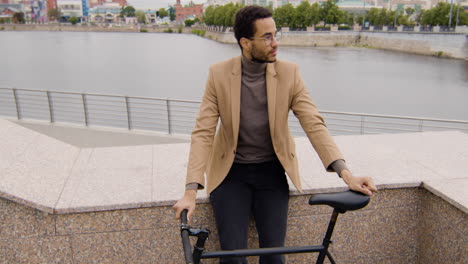 hombre americano sonriente mirando la cámara y sosteniendo su bicicleta mientras se apoya contra la pared en la ciudad 1
