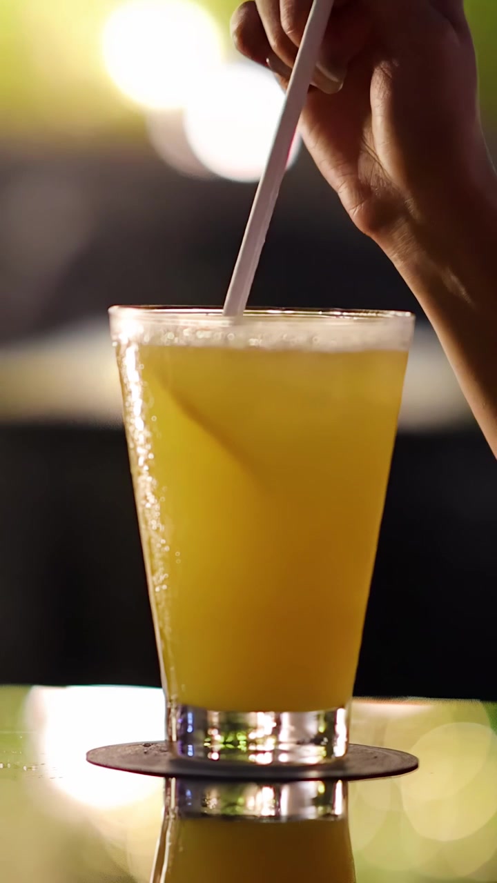 Yansıtıcı bir barda, pipetle sarı kokteyli karıştıran el, bokeh ışıkları, loş ışıklı akşam ambiyansı.