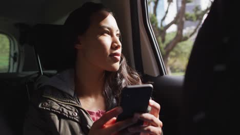 Mujer-Asiática-Sonriendo-Mientras-Mira-Por-La-Ventana-Y-Usa-Un-Teléfono-Inteligente-En-El-Auto