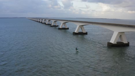 Aerial-backward-establishing-shot-of-a-Zeelandbrug-with-a-surfer-with-blue-wing-on-airfoil-under-it