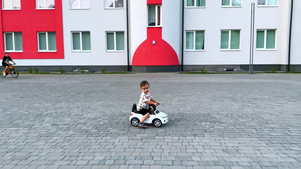 Un simpatico bambino piccolo che guida la sua auto giocattolo bianca all'aperto. Un bambino sorridente che si diverte attivamente in estate.
