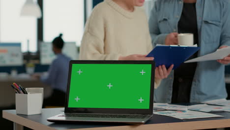laptop computer with green screen on desk in front of startup colleagues talking about sales charts papers