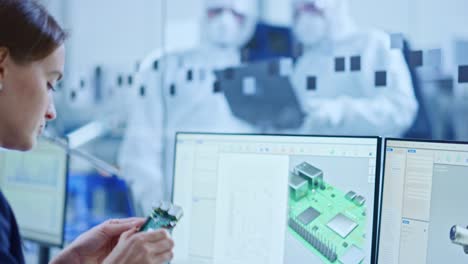 en taller limpio: trabajadores profesionales en trajes y máscaras usan computadora portátil y hablan. dentro de la oficina de la fábrica: ingeniera electrónica femenina trabaja en computadora, la pantalla muestra el diseño de placa de circuito impreso 3d