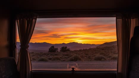 a tranquil sunset unfolds through a camper window, painting the sky with warm hues of orange, pink, and purple over majestic mountain ranges