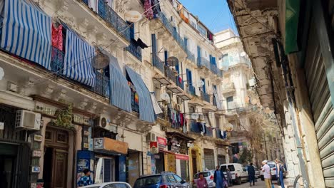 revealing shot on french colonial buildings in downtown algiers, locals, tourists and cars passing street, blue curtains hanging from windows on a sunny morning