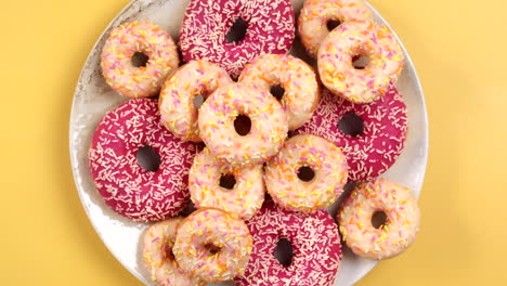 closeup of female hand taking small donut on plate full of colorful doughnuts in different size, pink and white on light yellow background. top view, filmed in prores.