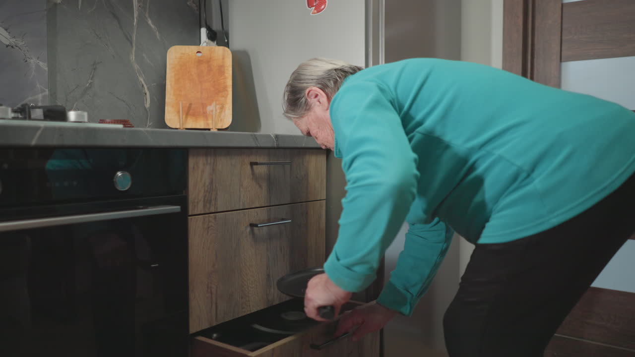 Elderly woman in green sweatshirt bending over to open kitchen drawer, searching for something. Modern kitchen with wooden accents and a marble countertop, capturing everyday routine in a home setting