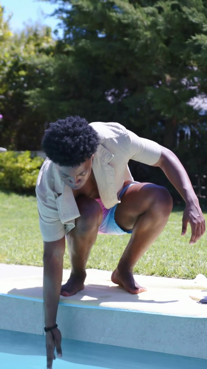 Vertical video: Glancing down man in swim shorts walking pool edge squatting testing water near hat